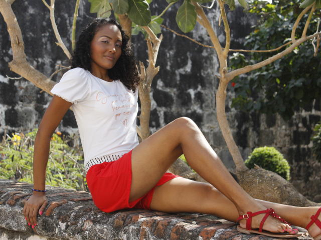 MeganGil, met een bruine huid en krullend zwart haar, draagt een witte blouse, rode shorts en rode sandalen. Ze zit op een bakstenen rand, met gekruiste benen, terwijl takken van een boom boven haar hangen. Ze glimlacht, ziet er ontspannen uit, en er schijnt zonlicht..