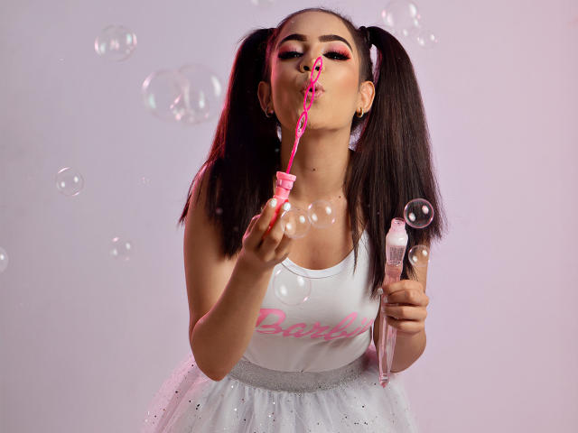 LunaRuiiz avec des couettes, une baguette à bulles rose, soufflant des bulles, un débardeur blanc, du rouge à lèvres rose, une jupe en tutu blanche, tenant une bouteille transparente, des yeux roses, une bulle dans la bouche, un fond rose et blanc..