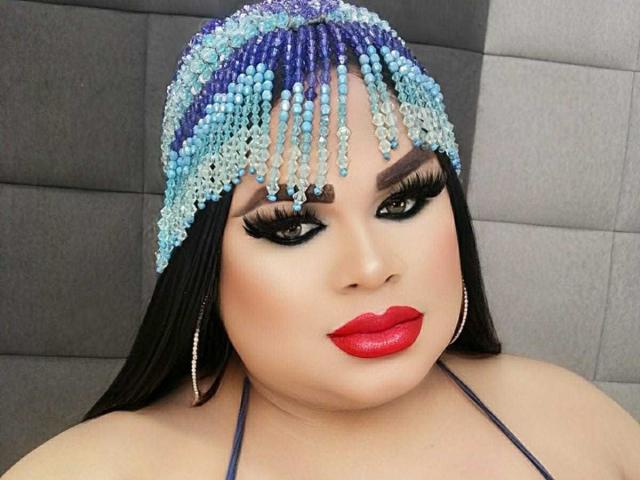 FabiolitaSex with big tits, wearing a blue and purple beaded headpiece, red lipstick, thick black eyebrows, and long black hair, against a grey tiled wall.