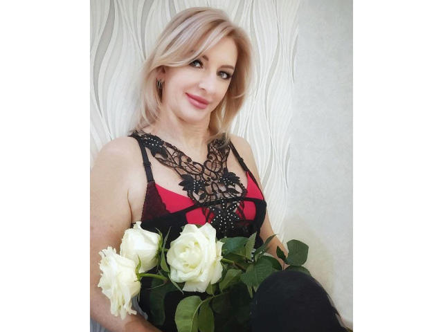 RoseQMagic con cabello rubio, blusa de encaje roja y negra, sosteniendo rosas blancas, sonriendo, usando pantalones negros, de pie frente a una pared blanca texturizada..