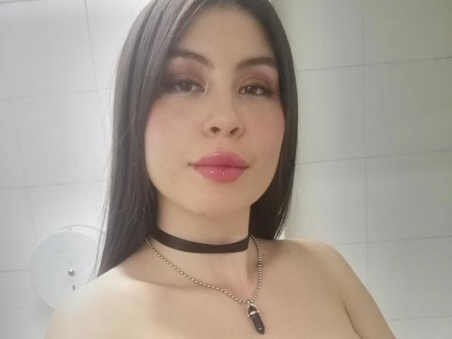 IsaTimeSweet ha lunghi capelli neri, rossetto rosa, una collana nera e una collana d'argento con una pietra viola. È senza top, con i seni visibili, davanti a un muro di piastrelle bianche..