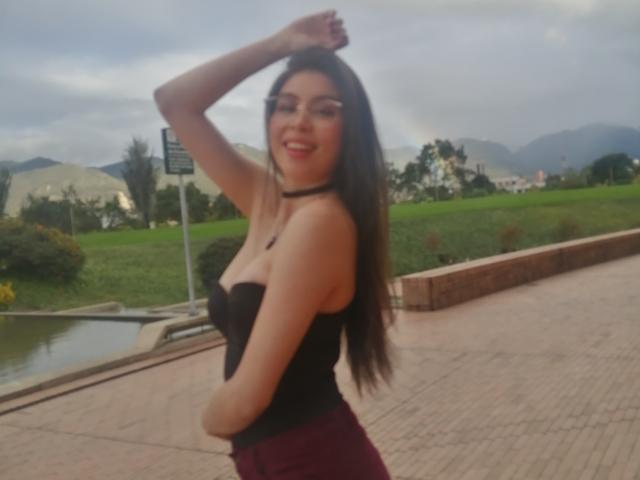 IsaTimeSweet, con lunghi capelli castani, sorridente, indossa un top nero senza spalline, pantaloni rossi e una collana nera, in posa con un braccio alzato, mostrando il seno, all'aperto in una giornata nuvolosa..