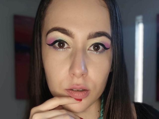 MabyLegrand has bright eye makeup with pink, green, and blue colors, and is biting her red-painted finger with her lips slightly parted. She has straight brown hair.