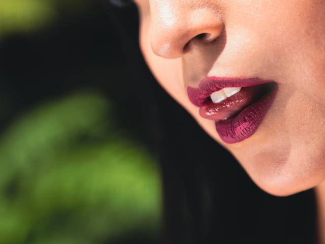 Los labios carnosos y brillantes de color magenta de ValentinaGomez están ligeramente entreabiertos, revelando un solo diente superior. Su piel lisa y bronceada está iluminada por la luz del sol. Su cabello negro es parcialmente visible..