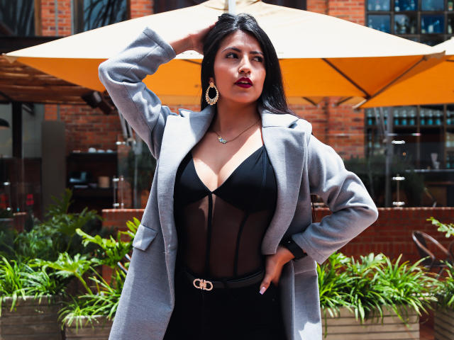 Valentina Gomez trägt ein schwarzes, transparentes Kleid, einen grauen Mantel, goldene Ohrringe und einen Gürtel. Sie hält ihre Hand im Mantel, steht unter einem gelben Regenschirm, im Freien bei Sonnenschein, umgeben von grünen Pflanzen, vor einem braunen Backsteinhaus. Sie wirkt selbstbewusst und sexy, hat eine wohlgeformte Brust..