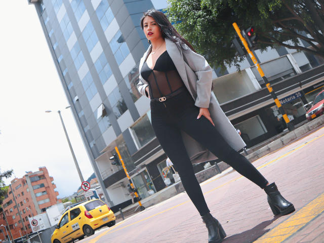 Valentina Gomez trägt eine schwarze, durchsichtige Bluse, einen grauen Mantel, schwarze Hosen und Stiefel. Sie steht mit den Händen in den Hüften, im Hintergrund ist ein gelbes Taxi zu sehen. Die Umgebung ist urban. Ihre Brüste sind sichtbar. Sie wirkt selbstbewusst und sexy..