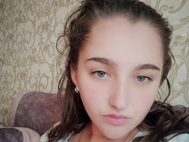 Lovelyy has light green eyes, brown wavy hair, and a small nose with pink lips. She's wearing flower earrings and a white shirt, looking directly at the camera.