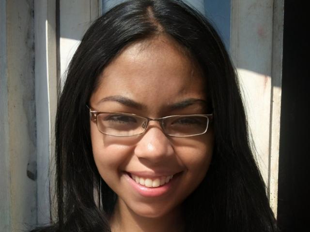 PerlaSexiy tiene la piel de color marrón oscuro, cabello negro largo y está sonriendo con gafas puestas. Se pueden ver sus pequeños senos y su nariz linda. Está mirando hacia abajo, con la luz del sol en su rostro..