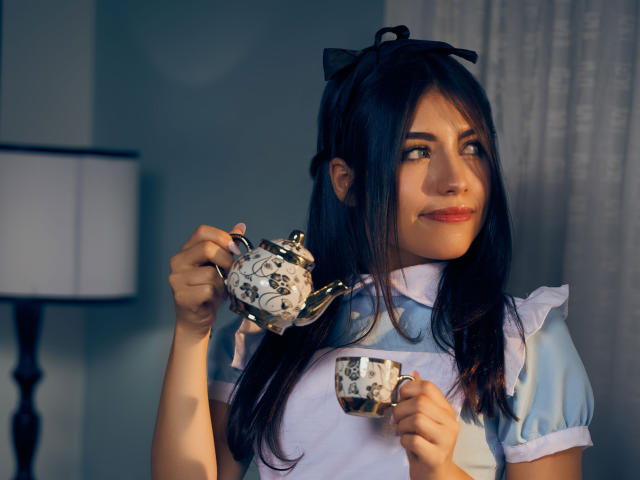 VanityValos mit dunklem Haar, das zu einem schwarzen Schleifchen gebunden ist, trägt ein blau-weißes Dienstmädchenkostüm und hält eine Teekanne mit Blumenmuster und eine passende Tasse. Sie blickt zur Seite und lächelt leicht..