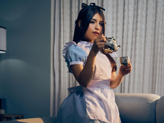 VanityValos con cabello castaño, vestida con un uniforme de sirvienta azul y blanco, sosteniendo una tetera y una taza, sentada en un sofá gris, y usando una cinta para el cabello negra..