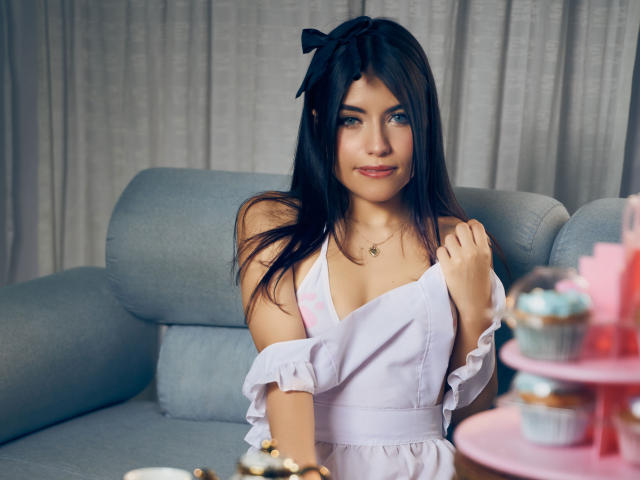 VanityValos with dark hair, blue eyes, and pink dress, showing small tits, smiling, hand on shoulder, gray couch, pink cupcake stand, white curtains. Soft, pretty, cute.