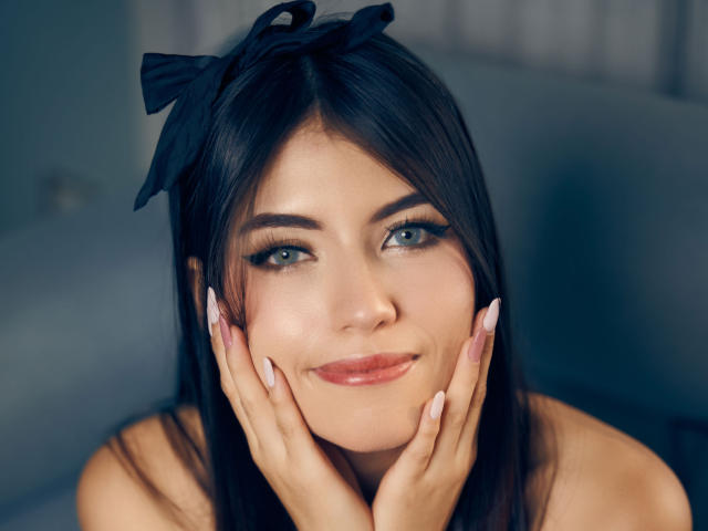 VanityValos has dark hair, blue eyes, and is smiling with hands on her face, showing off her pink nails. She's wearing a black bow in her hair.