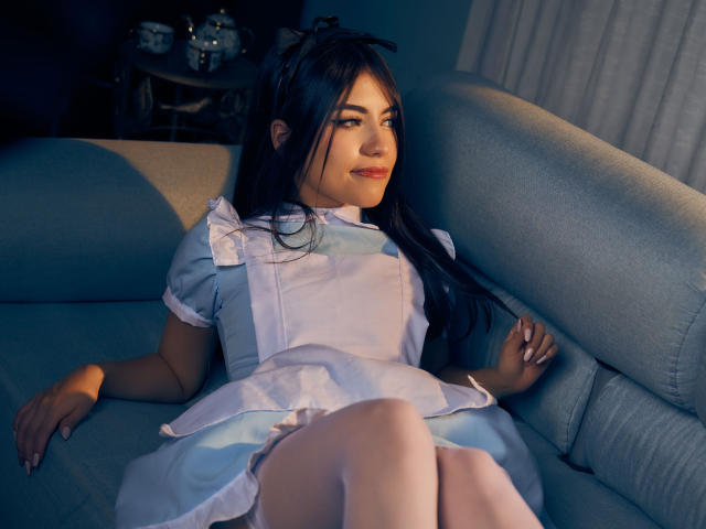 VanityValos, con cabello negro largo, vestida con un uniforme de sirvienta azul y blanco, sentada en un sofá gris, con las piernas cruzadas, mirando hacia un lado con una sonrisa sutil, luz suave..