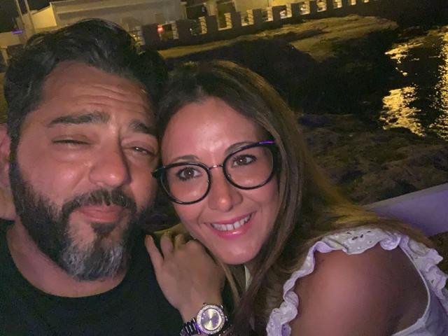 HotHellCouple, una coppia con occhiali e un sorriso, con il seno visibile sotto una camicetta bianca con volant, accanto a un uomo barbuto con una barba grigia, che fa l'occhiolino, in un ambiente poco illuminato..