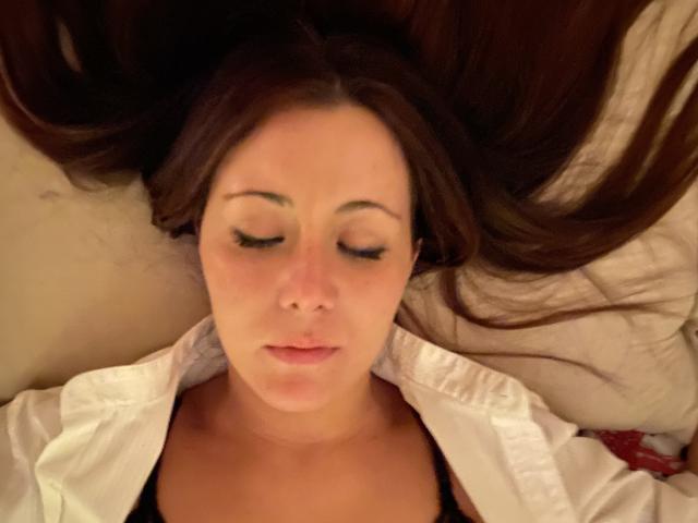 Il viso di HotHellCouple, occhi chiusi, lunghi capelli castani sparsi, camicia bianca aperta, top nero visibile, labbra rosa pallido, pelle leggermente arrossata, sdraiata, espressione rilassata..