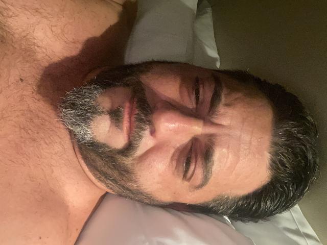 Coppia in pose sensuale, con barba e capelli scuri, occhi chiusi, sdraiata su un cuscino, con il petto visibile, che appare rilassata, leggera ombra sul viso, pelle media, leggera peluria sul petto..