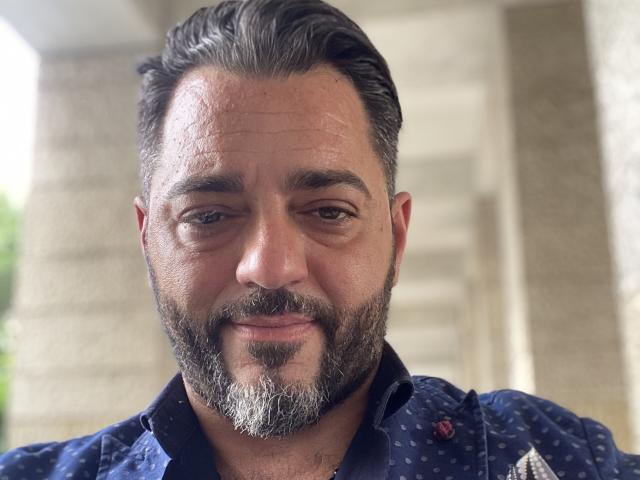HotHellCouple ha un leggero sorriso, una barba grigia e capelli corti, indossa una camicia blu a pois, con un viso rilassato, leggermente segnato dalle rughe, e occhi marroni dolci..
