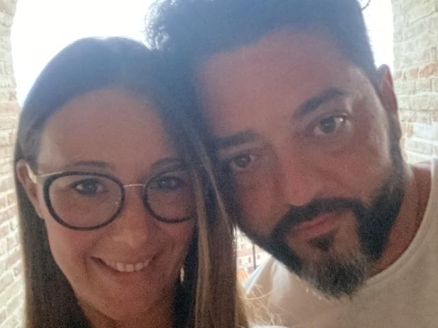 HotHellCouple con occhiali e capelli castani, che sorridono. HotHellCouple con barba scura e capelli corti, che sorride leggermente. Entrambi hanno la pelle chiara e indossano una camicia bianca..