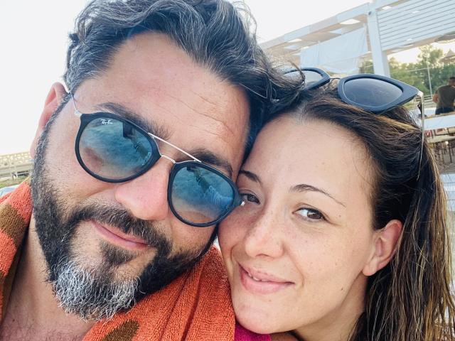 HotHellCouple mostra un uomo con barba e capelli grigi, occhiali da aviatore e un asciugamano arancione, accanto a una donna castana con lentiggini, che sorride e indossa gli occhiali sulla testa..