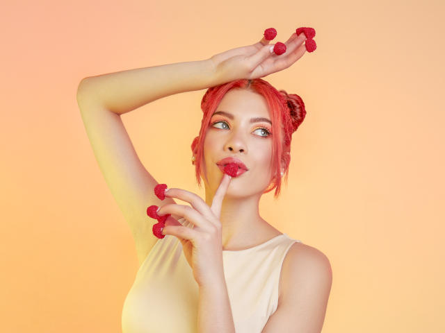 AshleyPort with red hair, wearing a white top, holding red raspberries, one in mouth, other in hand, arm raised, looking up, soft gradient background.