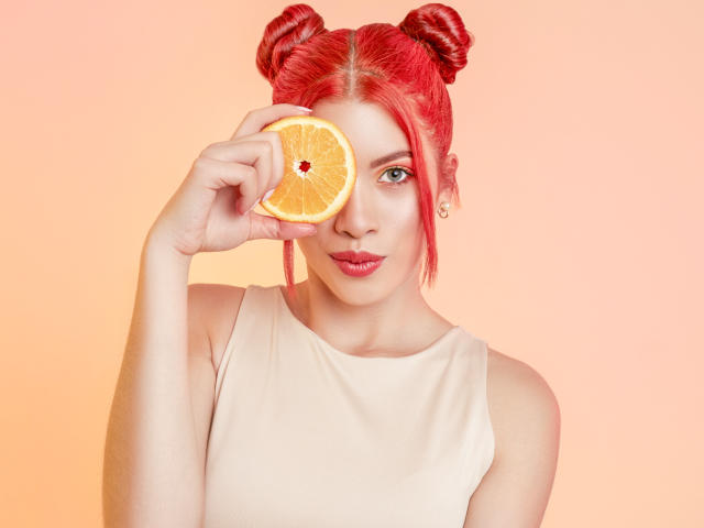 AshleyPort com cabelo ruivo em dois coques, segurando uma fatia de laranja sobre o olho direito, vestindo uma blusa bege sem mangas e um brinco de ouro, em um fundo cor de pêssego..