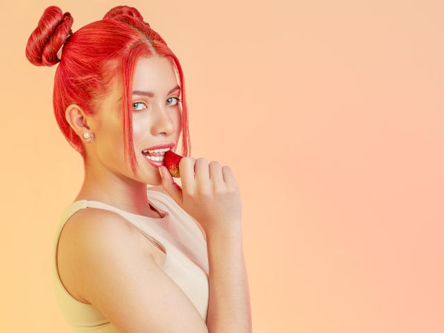 AshleyPort com cabelo ruivo vibrante preso em coques, sorrindo enquanto morde um pirulito vermelho, vestindo uma blusa branca sem mangas, segurando o pirulito perto da boca, fundo rosa degradê..