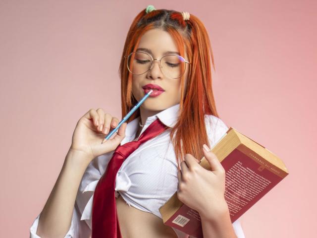 AshleyPort, met oranje haar, een bril en roze lippenstift, zuigt aan een blauwe pen terwijl ze een boek vasthoudt, ze draagt een witte blouse en een rode das, haar borsten zijn licht zichtbaar..