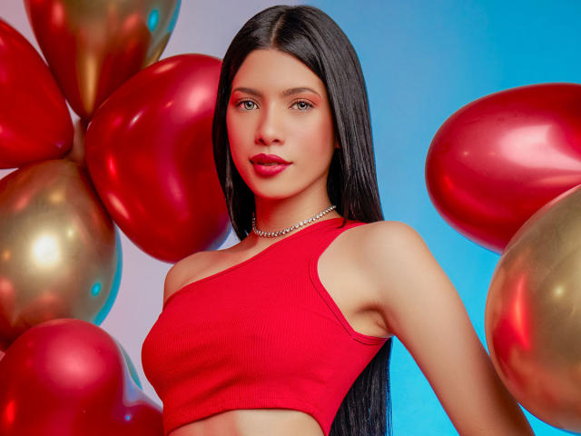 AshleyPort met lang zwart haar, een rode topje, waarbij haar borsten zichtbaar zijn, rode en gouden ballonnen, een blauwe achtergrond, draagt een zilveren ketting, kijkt naar de camera met rode lippenstift..