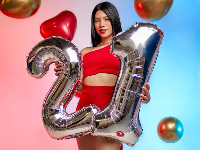 AshleyPort met een glanzende zilveren "2" ballon, een rode topje en een rode broek met hoge taille, die een ballon vasthoudt, met zwart haar, rode lippenstift, en gouden en rode ballonnen op de achtergrond..