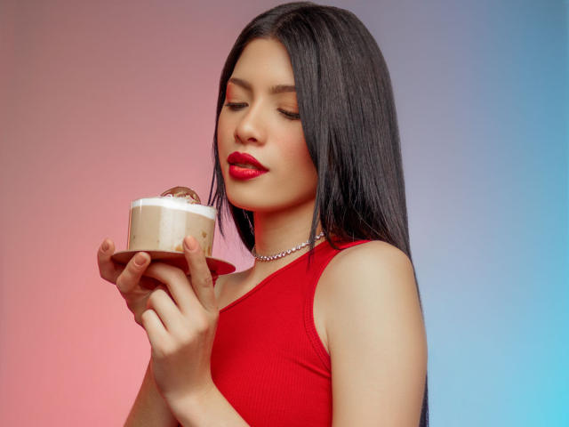 AshleyPort, con cabello negro largo, lápiz labial rojo y una blusa roja sin mangas, sosteniendo un pequeño cupcake de chocolate con glaseado blanco, llevando un collar de plata, mirando hacia abajo..