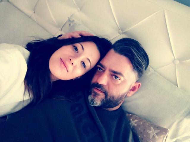 HotHellCouple, una ragazza con capelli scuri e labbra carnose, è sdraiata accanto a un uomo barbuto con i capelli pettinati all'indietro, entrambi che guardano la fotocamera, su un letto bianco imbottito..