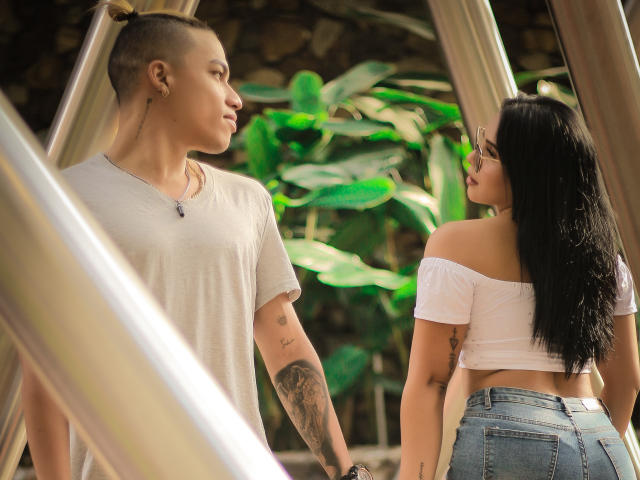 HoneyAndMaky with a topknot, wearing a white tee, looks at HoneyAndMaky with black hair, off-shoulder top, and blue jeans, standing in a sunlit, metal structure.