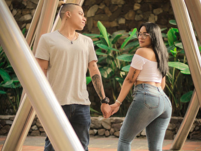 HoneyAndMaky, avec de longs cheveux noirs, des lunettes, un haut blanc à une épaule et un jean moulant, tenant la main d'un homme blond en t-shirt beige, se tient près de poutres métalliques..
