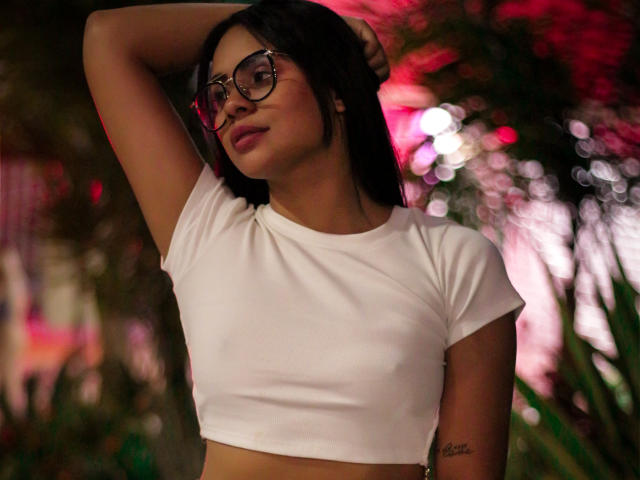 HoneyAndMaky usando óculos, com uma blusa cropped branca e braço levantado, mostrando uma pequena tatuagem no lado do corpo, com um fundo vermelho. O contorno dos seios é visível sob a blusa justa..