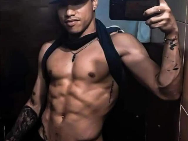 HoneyAndMaky, ohne Hemd, muskulös, gebräunte Haut, Tattoos am linken Arm, schwarze Kappe, schwarfer Schal, hält ein Telefon, Spiegelbild, definierte Bauchmuskeln, kleine Brustwarzen, leichtes Lächeln, rechter Arm angehoben..