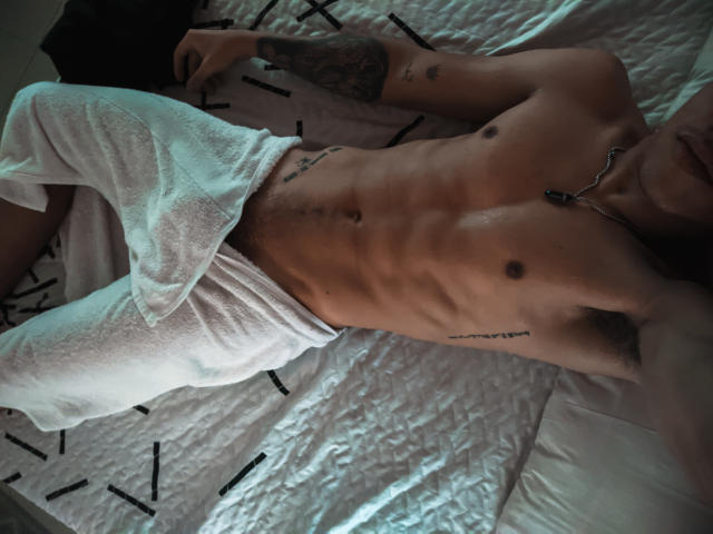 HoneyAndMaky lying on a bed, shirtless with white pants, showing a toned, hairy chest, tattoos, and a necklace. Their armpit and abs are visible.