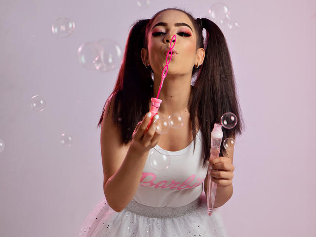 LunaRuiiz mit Zöpfen, aus denen ein rosa Seifenblase weht, in einem weißen Tanktop und einem weißen, glitzernden Kleid, hält einen rosa Seifenblasenstab, lutscht an einem rosa Faden, Seifenblasen schweben, rosa Lidschatten, helle Haut..