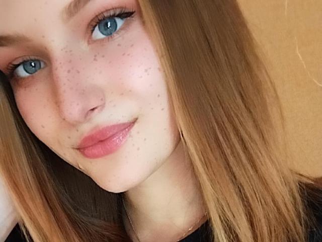 Het gezicht van SonyaNya, met haar blauwe ogen en lichte sproeten, staat in focus. Ze heeft steil, blond haar en roze lippen, en ze glimlacht zachtjes. Haar achterkant is niet zichtbaar op deze foto..