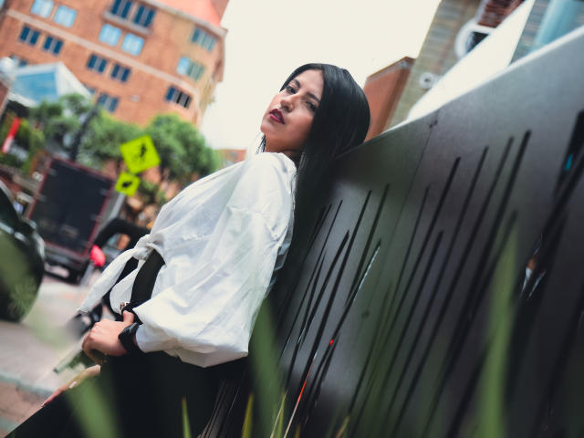 Valentina Gomez apoyada contra una pared de metal negro, vestida con una camisa blanca, pantalones negros y cabello largo y negro, con una mirada segura y ligeramente seductora, en un entorno urbano..