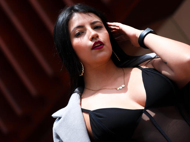 Valentina Gomez, con cabello negro, lápiz labial rojo oscuro, un sostén negro que deja ver sus pechos, una blusa transparente, una chaqueta gris, aretes dorados, un collar y una pulsera negra, mirando a la cámara..
