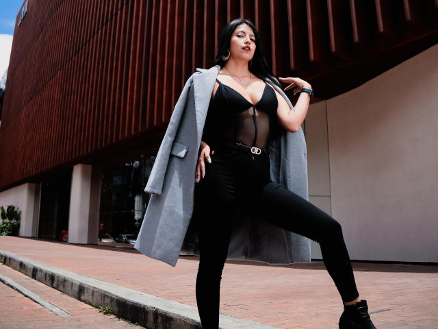 Valentina Gomez se mantiene erguida con confianza, vistiendo un abrigo gris sobre una blusa de malla negra que deja ver sus grandes senos, y pantalones negros ajustados con un cinturón, frente a un edificio oxidado..