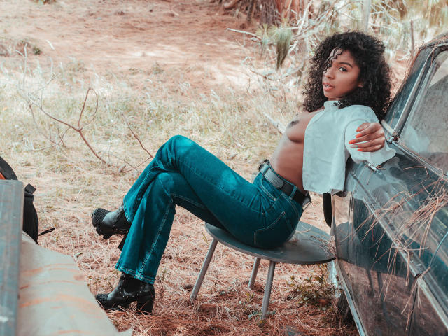 LisaMendez, topless, with curly black hair, blue jeans, black boots, and a white shirt, leaning on a car, showing her small, dark nipples and flat ass.