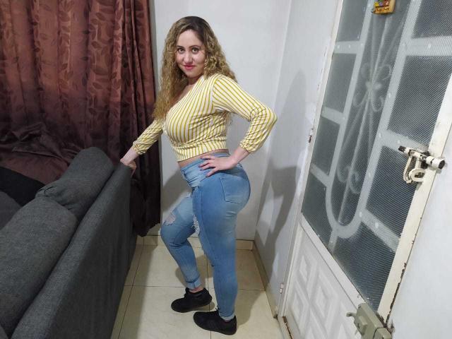 CamilaLovely è in piedi, con le mani sui fianchi, indossa una maglietta a righe gialle, jeans blu strappati e scarpe nere, mostrando i suoi fianchi formosi e il seno di media grandezza, in un soggiorno..