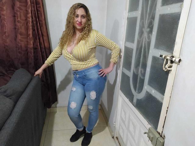 CamilaLovely, com cabelos loiros cacheados, camisa listrada amarela, decote profundo, jeans azuis rasgados, meias pretas, apoiada em um sofá cinza, porta branca e cortina marrom..