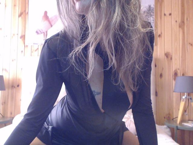 FrenchPlumeX, with wavy blonde hair, wearing a sheer black dress that reveals her tits and a small tattoo, leans forward in a wooden room.