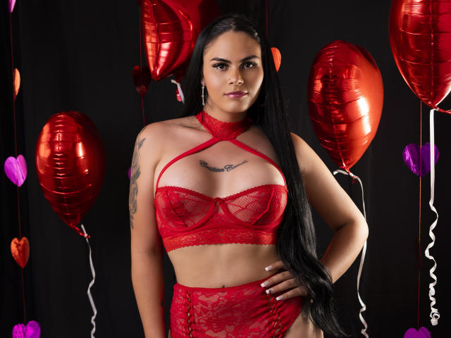 JassmineTS, com longos cabelos pretos, vestindo lingerie vermelha de renda, tem seios grandes, uma tatuagem no braço direito e balões vermelhos ao fundo. Ela está em pé com confiança, com a mão no quadril..