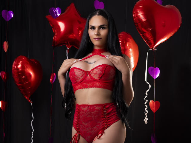 JassmineTS, avec ses longs cheveux noirs, portant de la lingerie en dentelle rouge, tenant ses seins, debout devant des ballons en forme de cœur rouges et violets, regardant l'appareil photo avec un léger sourire..