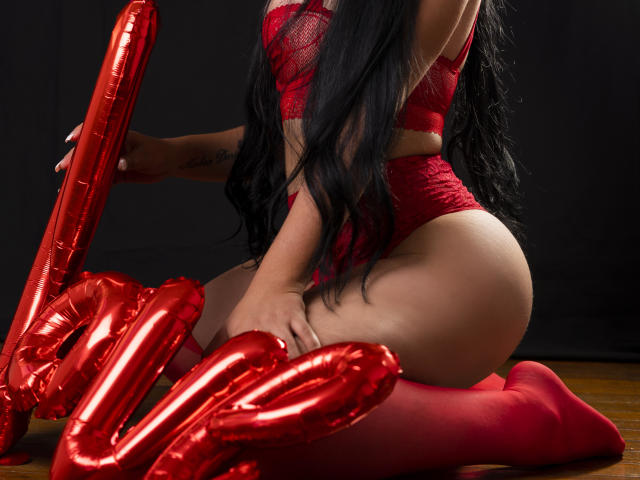 JassmineTS kneeling, red lace lingerie, red stockings, long black hair, red heart-shaped balloons, tattoo on left wrist, wooden floor, dark background, curvy butt, sexy, seductive pose.