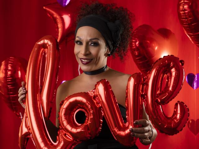 KhazandraX with curly afro, black headband, and choker, holding red "love" balloons, smiling with dark lipstick, wearing black strapless dress, red background.