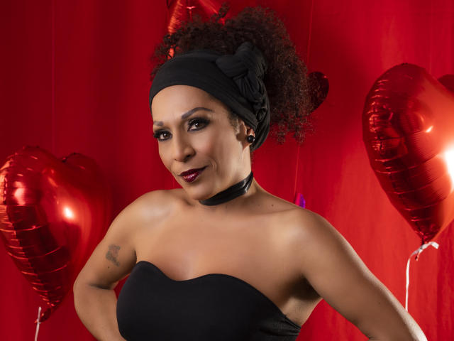 KhazandraX with curly black hair, black headband, black strapless top, choker, red lipstick, tattoo on left arm, red heart balloons, smiling, red background. Boobs visible, cute.